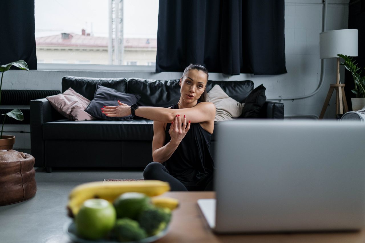Woman practicing yoga at home with a laptop, extending her arms in a warm and comfortable living room environment.
